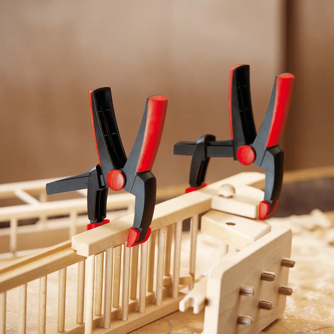 Zwei Kunststoffzwingen fixieren Holzbauteile. Deux pinces en plastique maintiennent des pièces en bois.